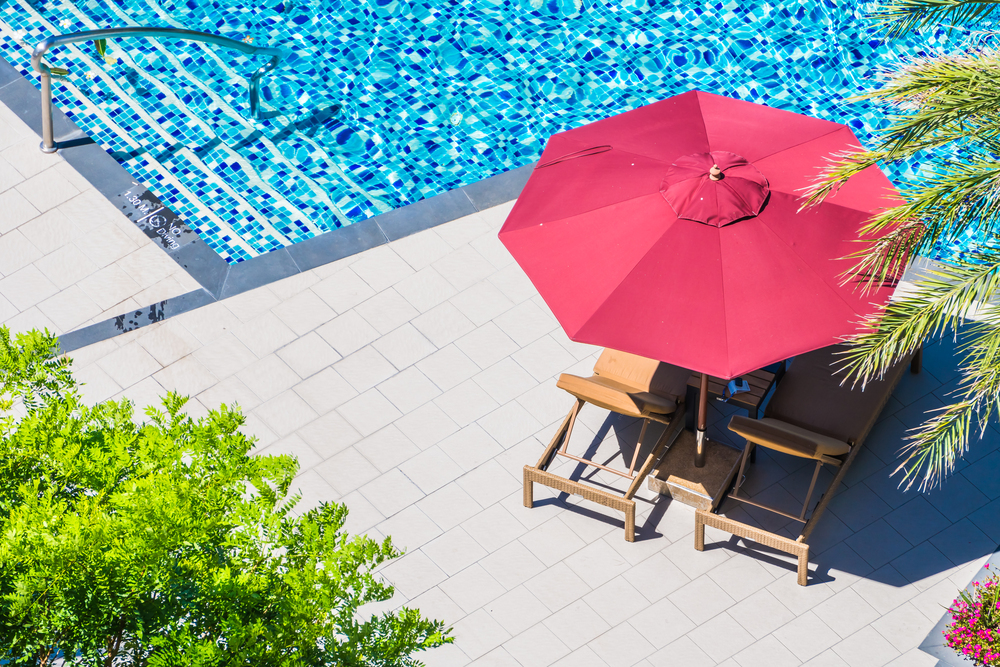 Área de descanso junto a una piscina, equipada con tumbonas y una sombrilla roja que brinda sombra, complementada por un entorno verde. La imagen refleja un espacio ideal para disfrutar de momentos al aire libre en días soleados.