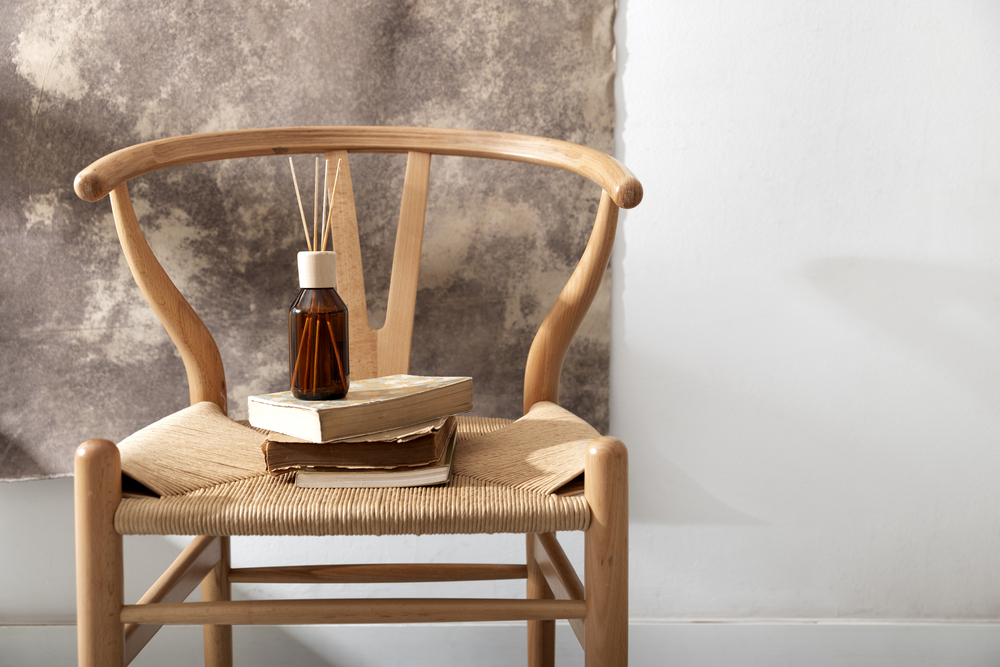 Silla de mimbre con un cojín blanco, junto a un difusor de fragancia y libros apilados, creando un rincón acogedor y natural en un ambiente decorativo.