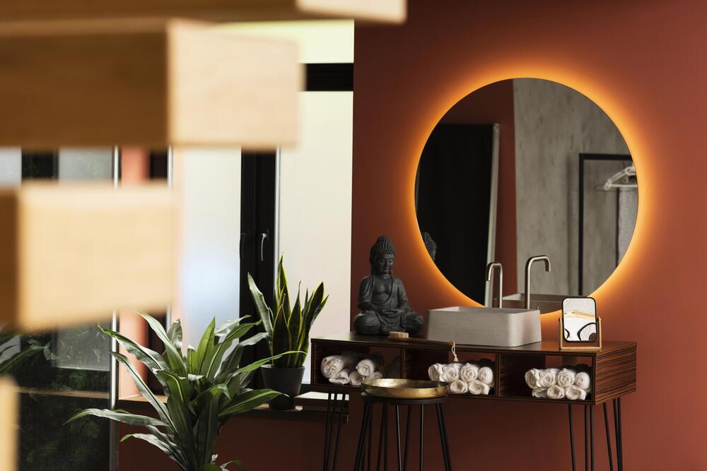 Baño contemporáneo con un espejo circular iluminado y un mueble de madera, complementado con una estatua de Buda y plantas, creando un espacio sereno y estético.