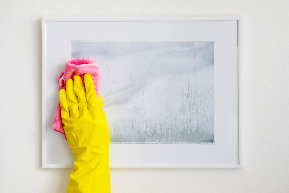 Una persona limpia el cristal de un cuadro con un paño rosa mientras usa guantes amarillos, asegurándose de mantener la obra de arte en perfectas condiciones. El cuadro está enmarcado con un marco blanco, reflejando un enfoque meticuloso hacia la conservación del arte.