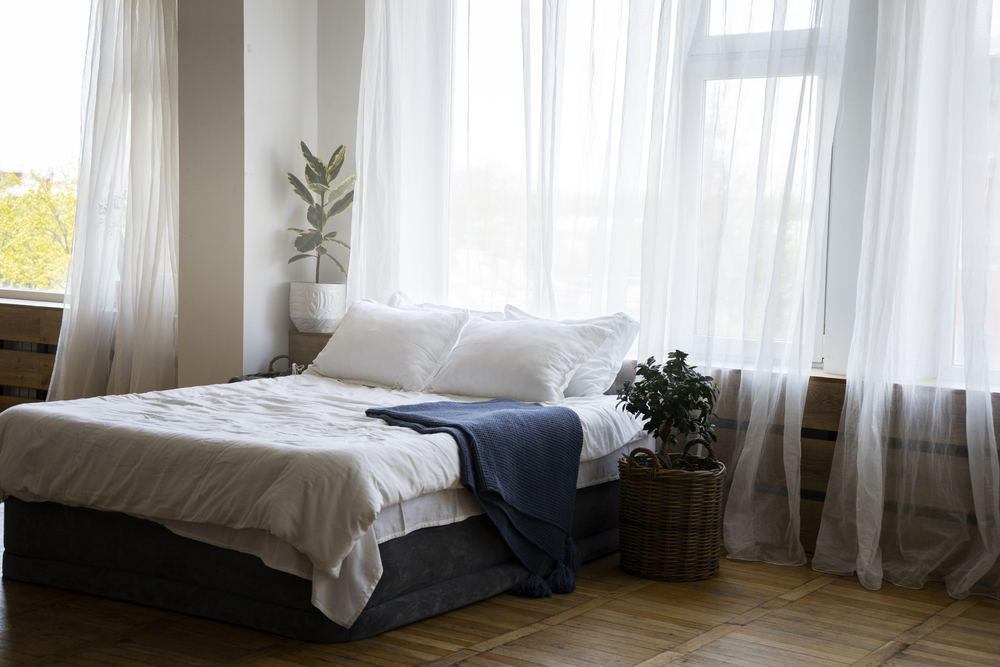 Esta habitación presenta una cama cómoda con sábanas blancas y una manta azul, rodeada de luz natural que entra por grandes ventanas. Las cortinas ligeras y las plantas aportan frescura y un ambiente relajante, ideal para un descanso reparador y un refugio personal.