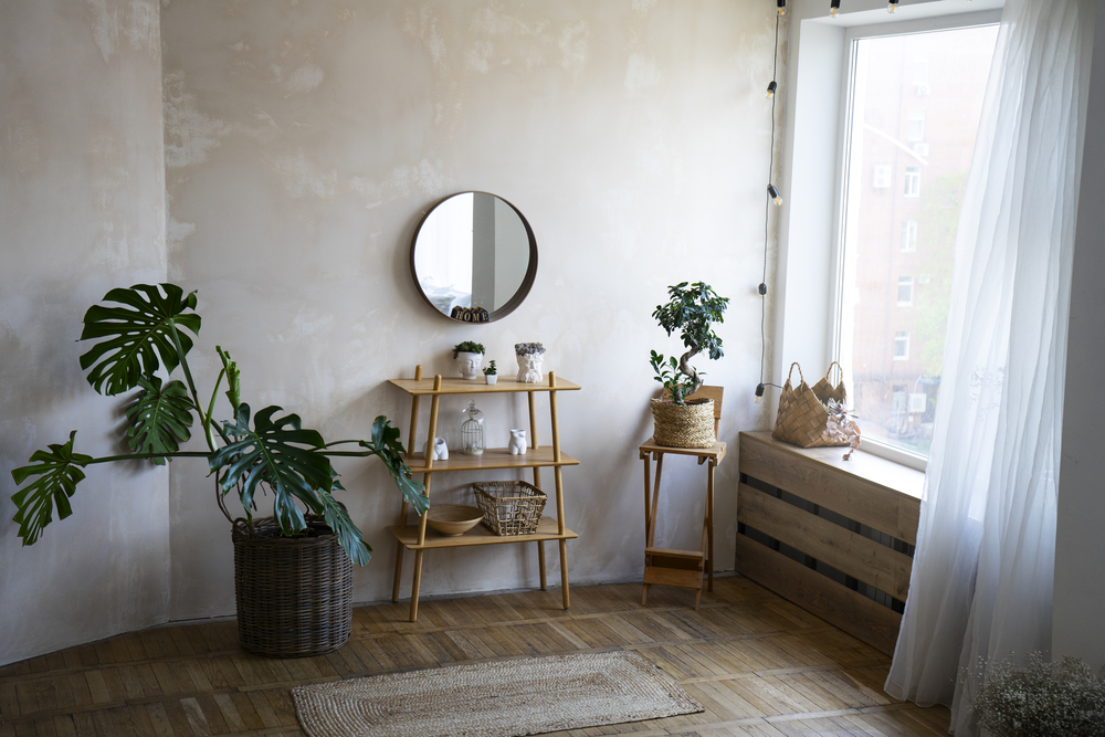 Esquina decorativa con un espejo circular que refleja luz natural, acompañado de una estantería de madera y plantas, creando un ambiente armonioso y relajante.