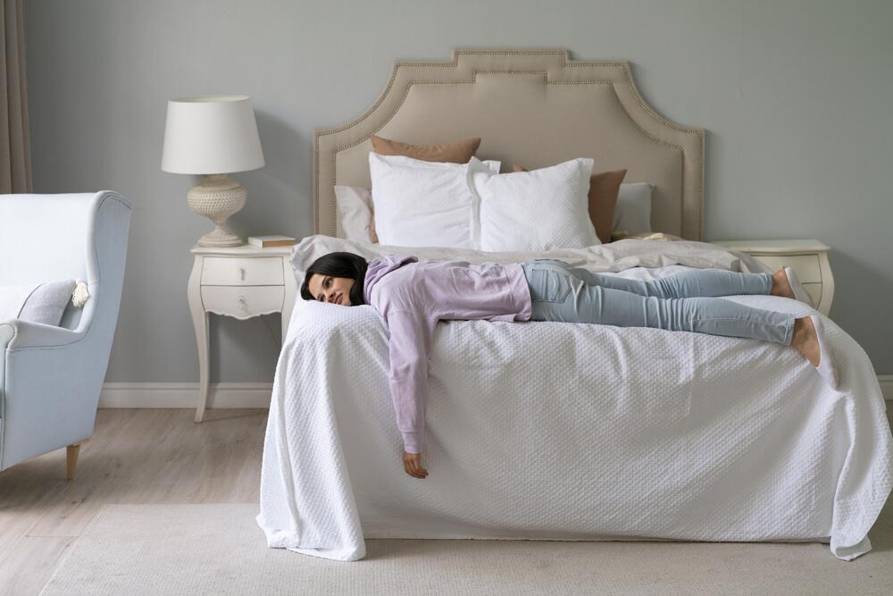 Una mujer se desploma sobre una cama con sábanas blancas y almohadas, mostrando la necesidad de descanso. La imagen ilustra la importancia de elegir la cama perfecta para un sueño reparador y un bienestar óptimo.