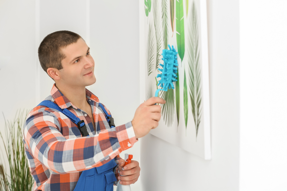 Un hombre está limpiando un cuadro decorativo en la pared utilizando un paño azul y un spray limpiador. La imagen ilustra la importancia del cuidado y la limpieza adecuada de las obras de arte para preservar su belleza y longevidad.