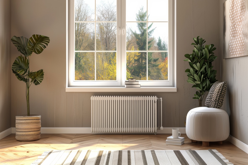 Un radiador blanco se encuentra en un ambiente cálido y acogedor, situado debajo de una ventana que permite ver un hermoso paisaje otoñal. El espacio incluye plantas en macetas y un asiento cómodo, creando una atmósfera ideal para disfrutar del confort del hogar.